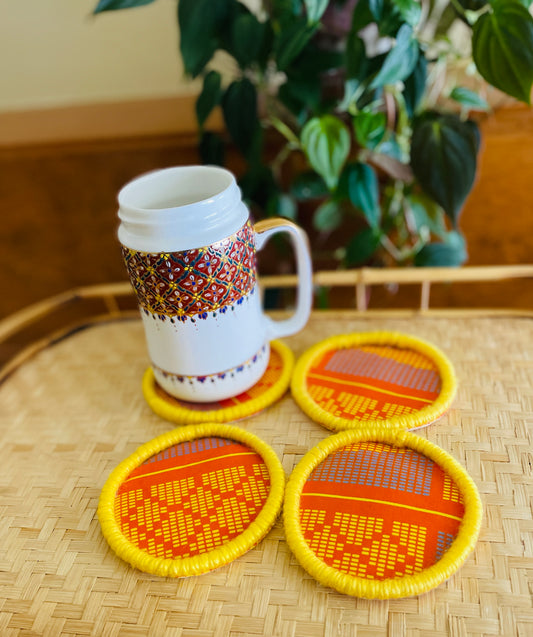 orange and yellow print coaster set Made with African wax fabric with a yellow border made of yarn wrapped rope with felt fabric on the bottom to help absorb moisture.  Materials:fabric, felt fabric, rope, yarn  Dimensions: