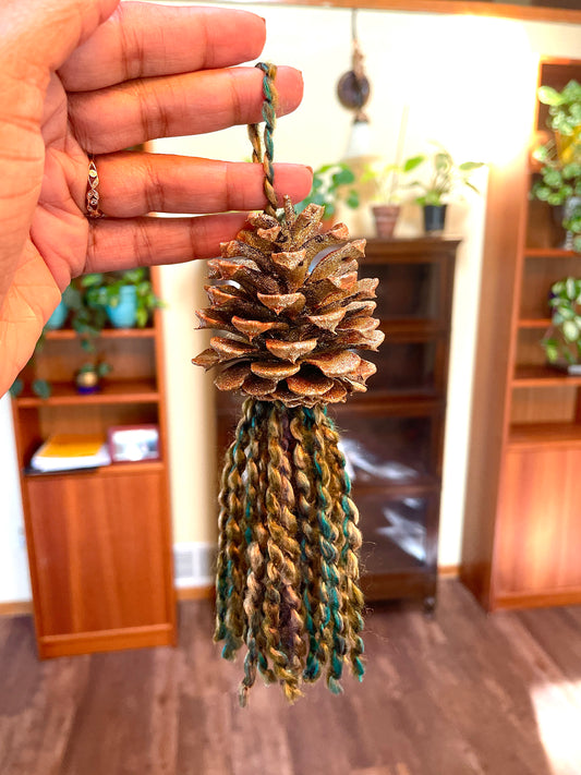 Pine cone sprayed with bronze paint and multicolor shimmery yarn with gold and green fringe  Materials: pine cone, spray paint, yarn