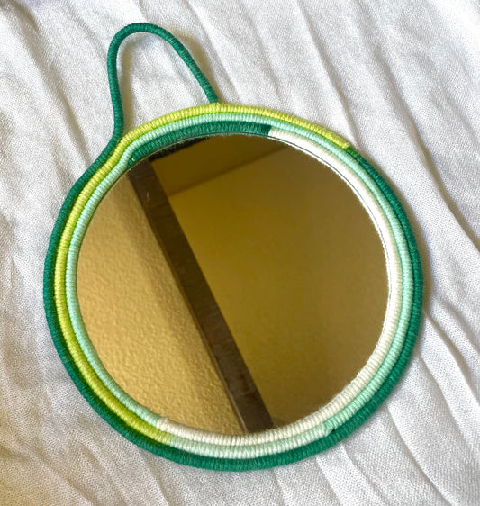 Lime green, white, sage and mint green yarn wrapped around rope on the frame on an 8 inch mirror with strap for hanging  Dimensions: 12.5x9.5 inches  Materials: 8 inch round mirror, yarn, felt fabric