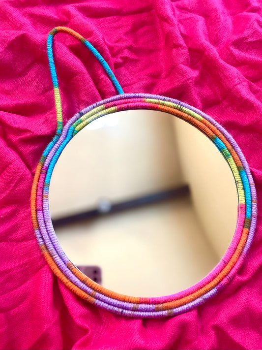 Multicolored (blue, pink, lavender, orange, yellow) yarn wrapped around rope on the frame on an 8 inch mirror with strap for hanging  Dimensions: 12.5x9.5 inches  Materials: 8 inch round mirror, yarn, felt fabric