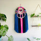 pink blue black white wall hanging Made with pink, blue, black and white yarn with upper center yarn wrapping detail and pompom and has 2 tiers of fringe.  Materials: yarn, bamboo hoops  Dimensions: 30 in L x 12 W 