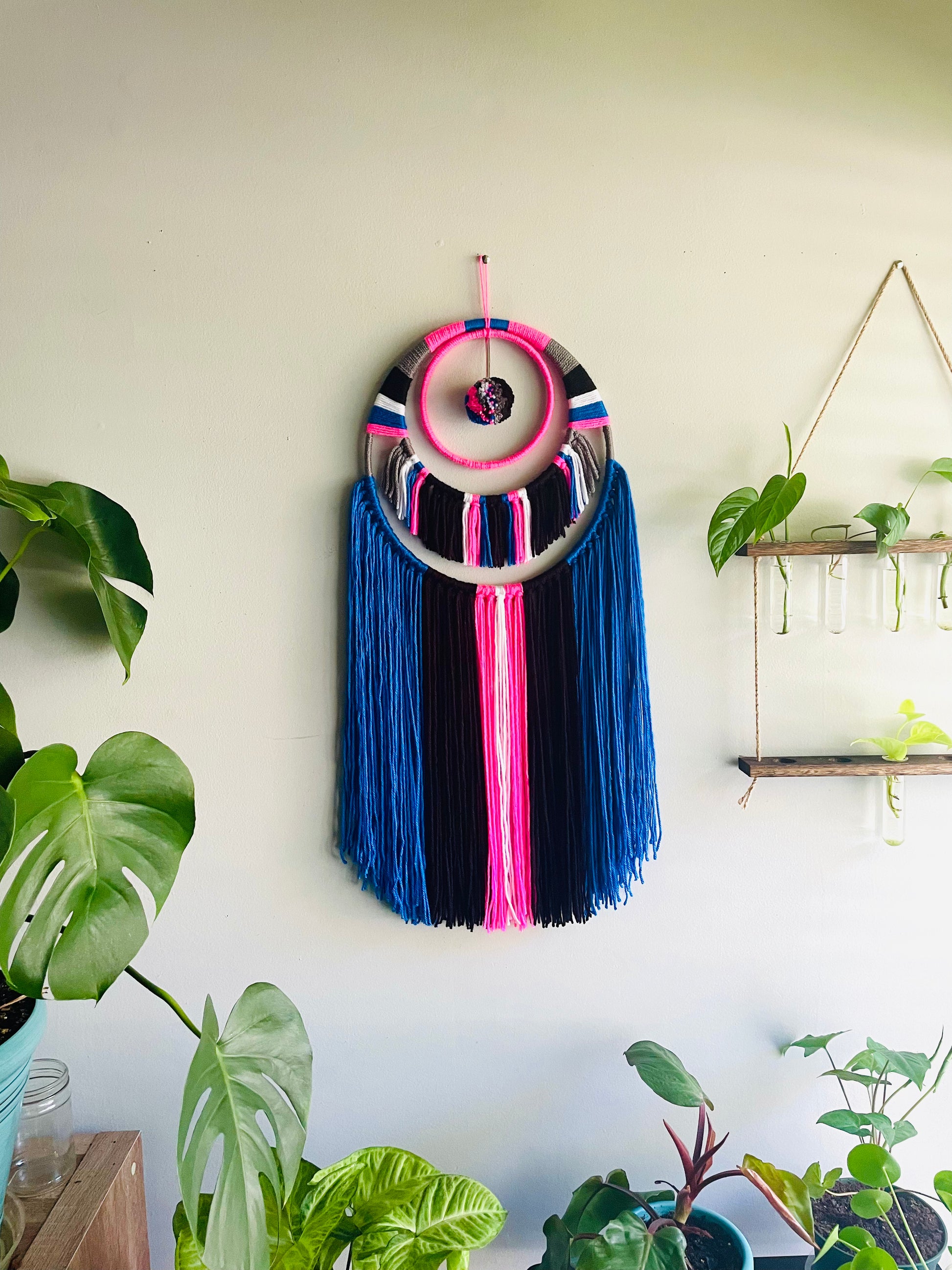 pink blue black white wall hanging Made with pink, blue, black and white yarn with upper center yarn wrapping detail and pompom and has 2 tiers of fringe.  Materials: yarn, bamboo hoops  Dimensions: 30 in L x 12 W 